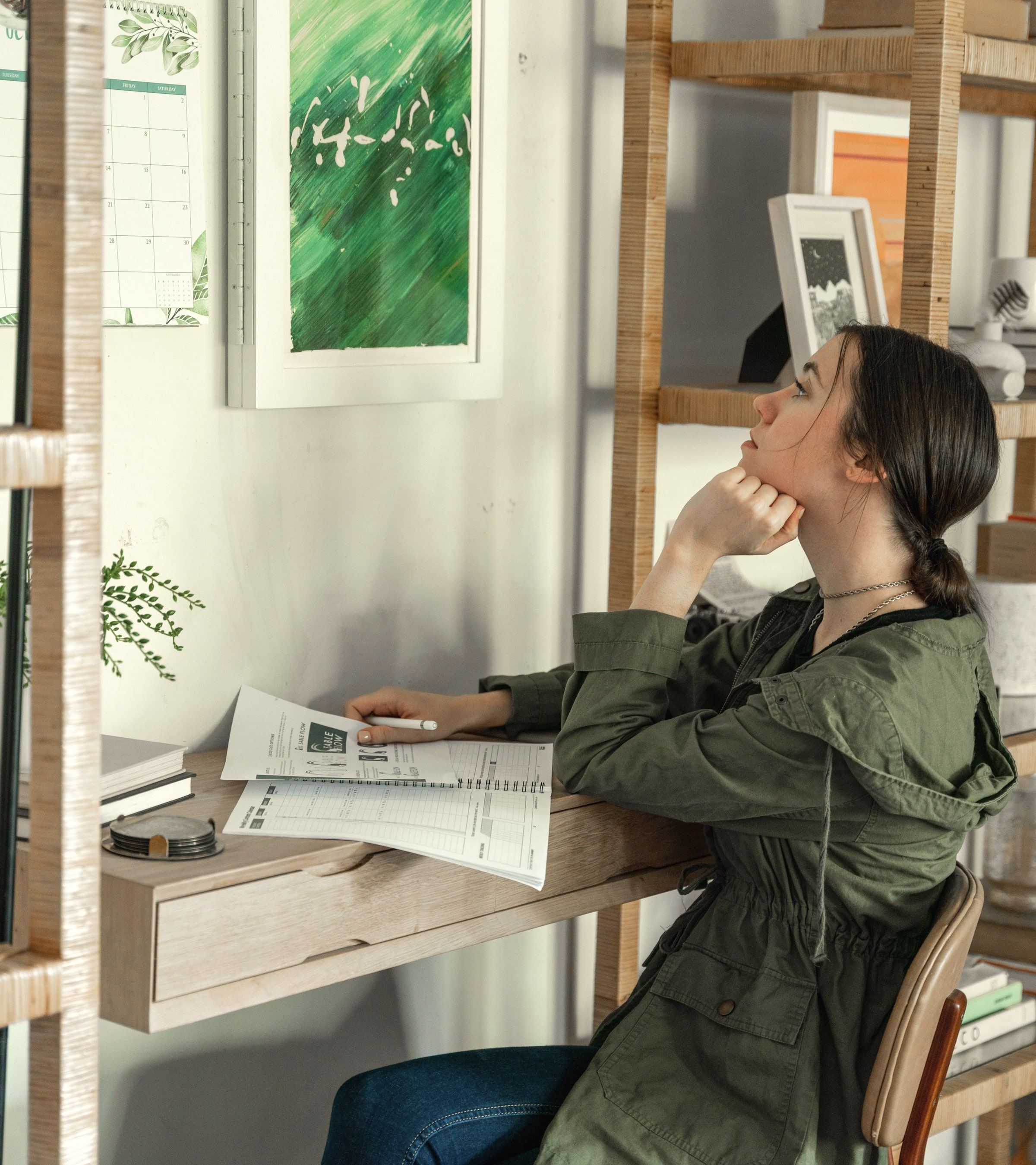 A student sits at a desk with papers, looking up in thought.