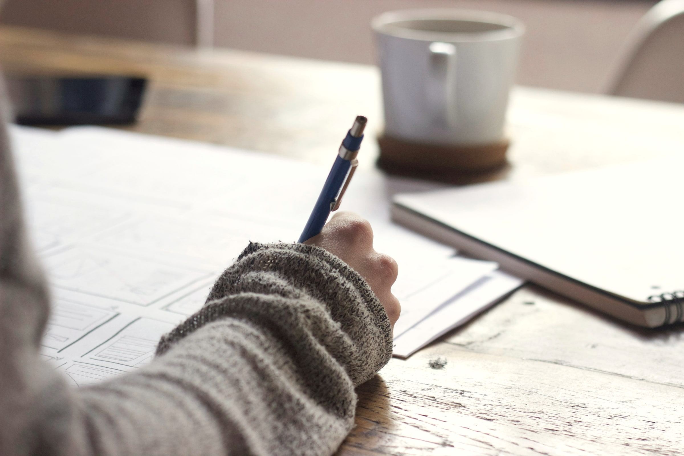 A student's hand writing notes on paper beside a mug on a wooden table.